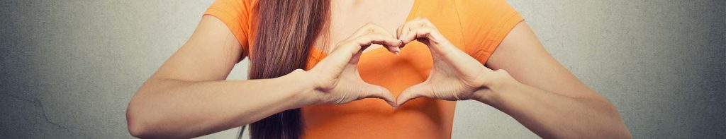 Nahaufnahme vom Oberkörper einer Frau in orange farbenem T-Shirt, die mit ihren Händen ein Herz über ihrem Herzen formt