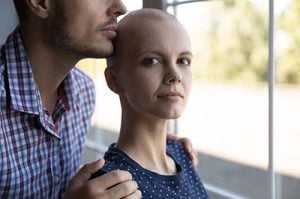 Shot of a young couple, the man stands protectively behind the young bald woman, who you can see has just undergone chemotherapy