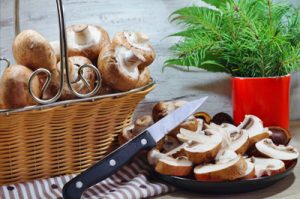 On voit un panier en osier avec des champignons bruns frais et une assiette de champignons émincés, avec un pot d'herbes aromatiques en arrière-plan.