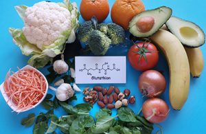 Des légumes et des fruits sains sur fond bleu, entre lesquels se trouve une fiche blanche avec la formule chimique du gluthathion.