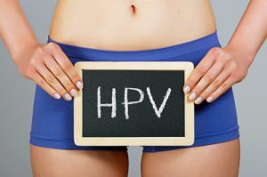 Grande plano de uma jovem mulher, em roupa interior azul, segurando uma pastilha com as letras HPV na sua frente.