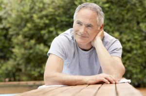 Mann im Seniorenalter, der draußen an einem Holztisch sitzt und zufrieden lächelnd in die Ferne blickt