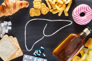 On a black table in the center is drawn with chalk the typical outline of a liver. Around it are draped unhealthy food, alcohol and drugs