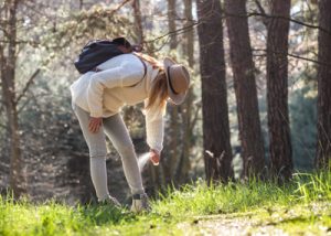 Eine Frau in Wanderkleidung steht im Wald und sprüht sich Zeckenschutzmittel an ihre Waden