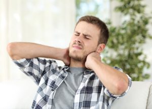Shot of young man in plaid shirt sitting on sofa grabbing his aching neck