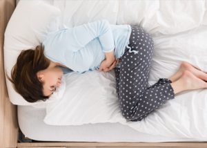 Fotografia de uma jovem deitada com cãibras na cama segurando a zona do estômago com ambas as mãos